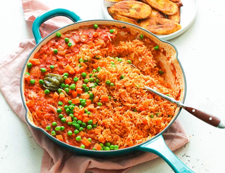 Jollof Rice in a Skillet with peas and fried sweet plantains on a white plate.